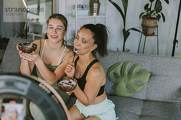 Weibliche Fitnesstrainerinnen essen Obstschalen  während sie zu Hause Vlogging betreiben