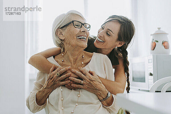 Fröhliche Frau  die ihre in der Küche sitzende Großmutter umarmt