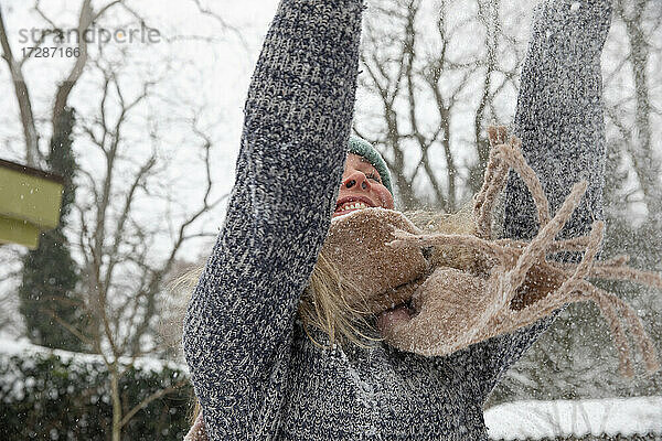 Glückliche reife Frau mit erhobenen Armen  die Schnee wirft  während sie im Hinterhof spielt