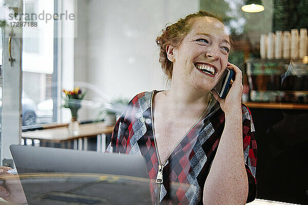 Glückliche Frau im mittleren Erwachsenenalter  die wegschaut  während sie in einem Café mit einem Mobiltelefon spricht