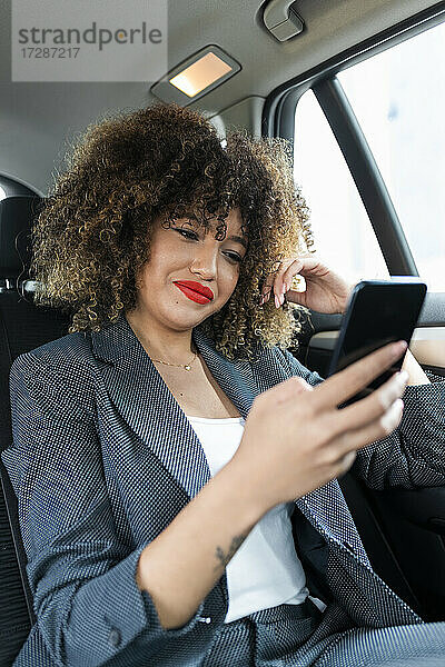 Weibliche Fachkraft mit lockigem Haar  die im Auto sitzend ein Mobiltelefon benutzt
