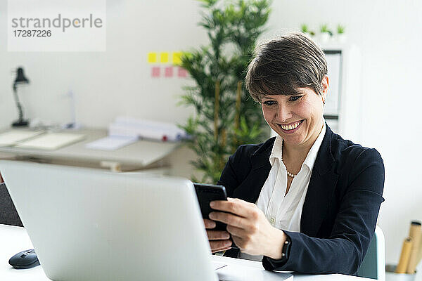 Fröhliche weibliche professionelle Textnachrichten über ein Smartphone  während sie am Schreibtisch im Büro sitzt