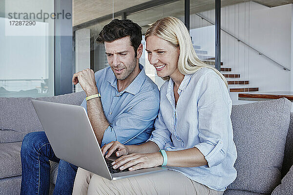 Lächelnde blonde Frau  die einen Laptop benutzt  während ein Mann auf dem Sofa im Wohnzimmer sitzt