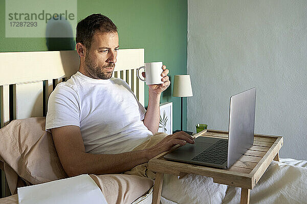 Mittlerer erwachsener Mann mit Kaffeetasse und Laptop  während er sich im Schlafzimmer ausruht
