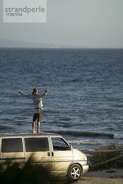 Junger Mann mit ausgestreckten Armen auf dem Dach eines Lieferwagens am Meer stehend