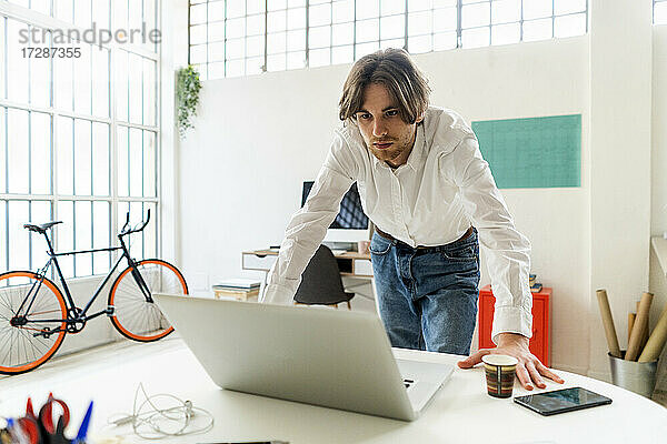 Geschäftsmann schaut auf einen Laptop und lehnt sich an einen Schreibtisch in einem kreativen Büro