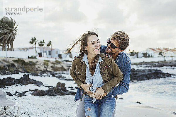 Lächelnder Mann umarmt Frau von hinten am Strand