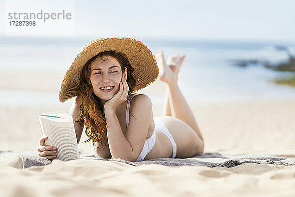 Lächelnde schöne Frau mit Buch Tag träumt  während auf Sand während sonnigen Tag liegen