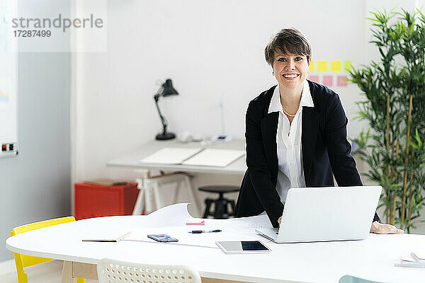 Lächelnde Geschäftsfrau mit Laptop am Schreibtisch im Büro stehend