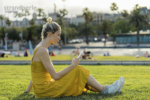 Frau  die im Gras sitzend eine SMS über ihr Mobiltelefon verschickt