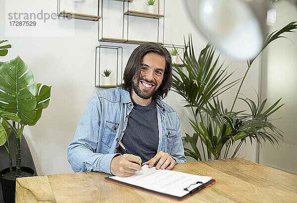 Lächelnder Geschäftsmann  der ein Dokument unterzeichnet  während er am Schreibtisch im Büro sitzt