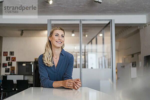 Lächelnde weibliche Fachkraft  die wegschaut und sich auf einen Schreibtisch im Büro stützt