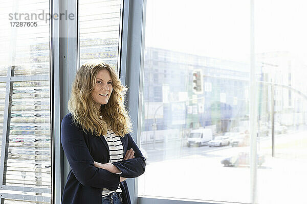 Lächelnde Unternehmerin schaut weg  während sie vor einem Glasfenster im Büro steht