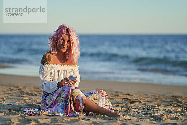 Frau mit rosafarbenen Haaren  die ihr Handy hält  während sie bei Sonnenuntergang am Strand sitzt