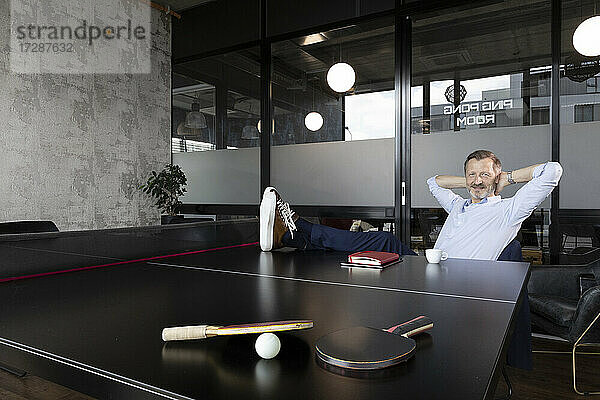Geschäftsmann mit Händen hinter dem Kopf sitzt an der Tischtennisplatte im Büro