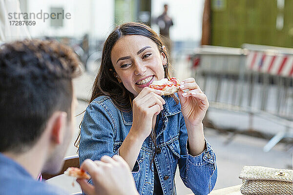 Junge Frau mit Pizzastück lächelt und sieht einen Mann an