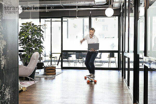 Lächelnde männliche Fachkraft mit Laptop fährt Skateboard im Büro