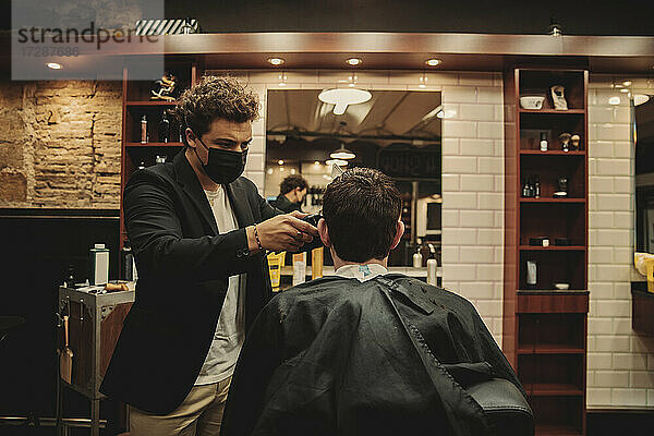 Männlicher Friseur mit Gesichtsschutzmaske beim Schneiden der Haare eines Kunden in einem Friseursalon