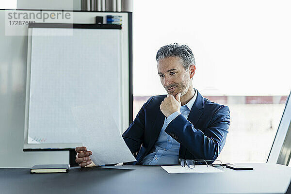 Älterer Geschäftsmann mit Hand am Kinn  der ein Dokument am Schreibtisch im Büro liest
