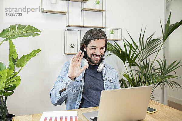 Lächelnde männliche Fachkraft winkt bei einem Videoanruf über einen Laptop im Büro