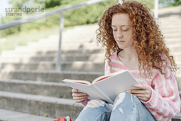 Rothaarige Frau mit lockigem Haar liest ein Buch  während sie auf einer Treppe sitzt