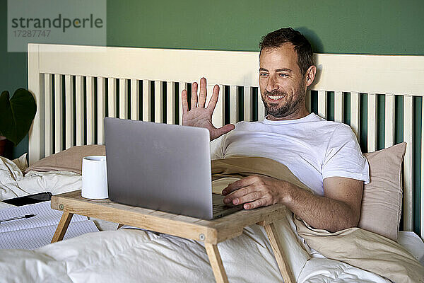 Lächelnder Mann winkt mit der Hand zu einem Videoanruf über einen Laptop zu Hause