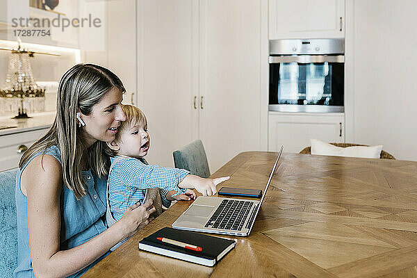 Junge zeigt auf Laptop  während er auf dem Schoß der Mutter sitzt und in der Küche arbeitet