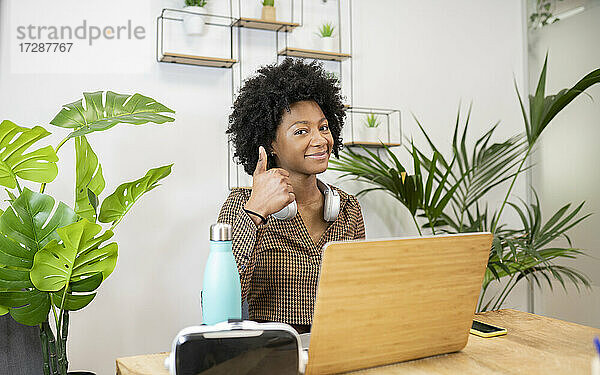 Lächelnde weibliche Fachkraft gestikuliert Daumen nach oben beim Sitzen am Schreibtisch im Büro