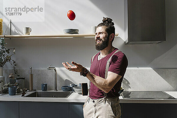 Bärtiger Mann spielt mit Tomate  während er an der Küchentheke steht