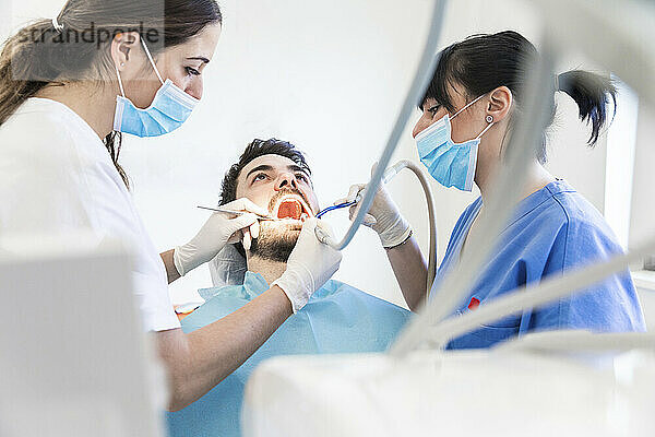 Weibliche Zahnärzte beim Bohren von Zähnen eines männlichen Patienten in einer Klinik während der Pandemie