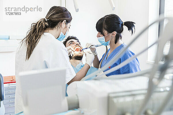 Zahnärztin mit Assistentin beim Bohren der Zähne eines männlichen Patienten in einer Klinik