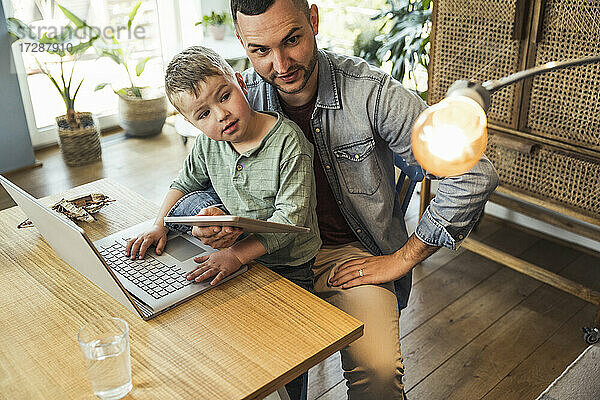 Vater und Sohn betrachten eine Glühbirne  während sie mit Tablet und Laptop in einem intelligenten Haus sitzen