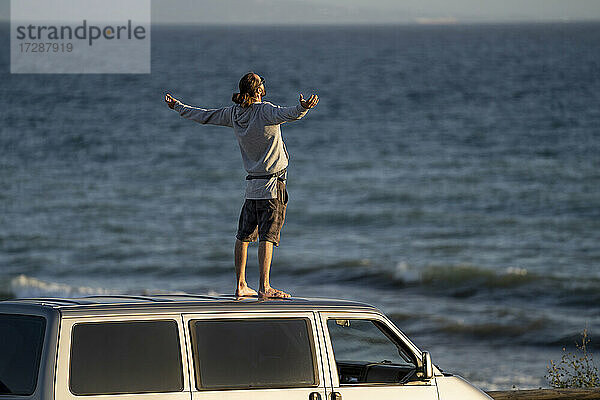 Unbekümmerter Mann steht mit ausgestreckten Armen auf dem Dach eines Minivans