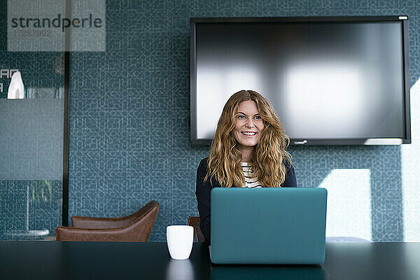 Lächelnde weibliche Fachkraft mit Laptop vor einem Fernseher im Büro sitzend
