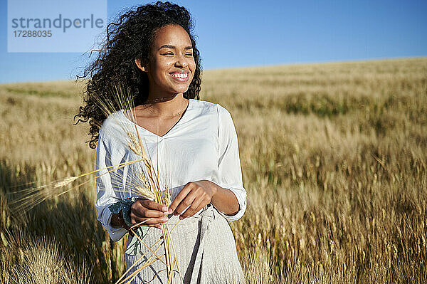 Lächelnde junge Frau hält Weizenernte auf einem Feld an einem sonnigen Tag