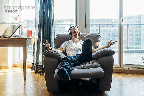 Lächelnder Mann mit Kopfhörern  der auf einem Lehnstuhl im Wohnzimmer sitzt und gestikuliert