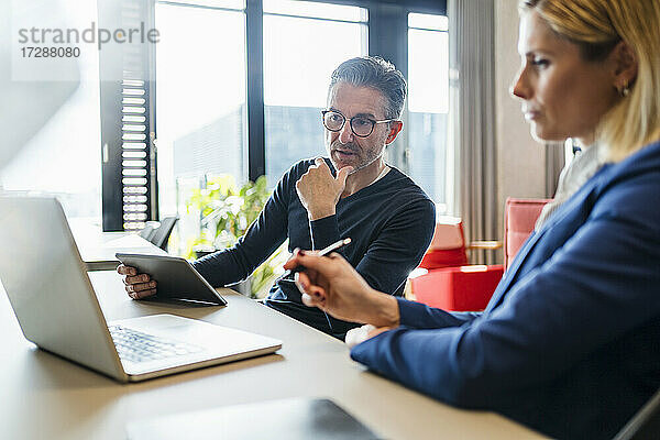 Älterer Geschäftsmann  der auf seinen Laptop schaut  während er mit einer Mitarbeiterin in einem kreativen Büro diskutiert