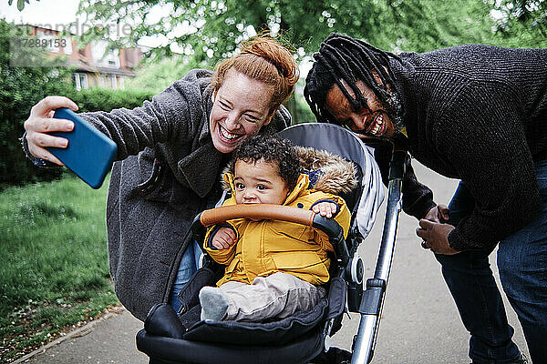 Fröhliche Eltern machen ein Selfie mit ihrem Sohn im Kinderwagen im Park