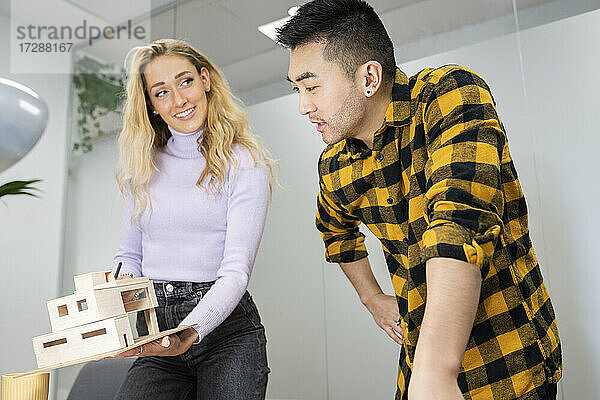 Junger Geschäftsmann im Gespräch mit einer Kollegin  die ein Modellhaus im Büro hält
