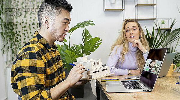 Männlicher Architekt  der ein Hausmodell per Videoanruf im Büro erklärt
