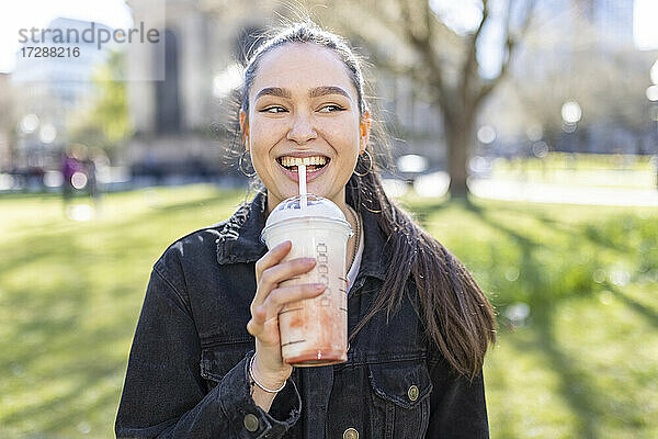 Lächelnde Frau  die wegschaut  während sie in einem öffentlichen Park einen Milchshake trinkt