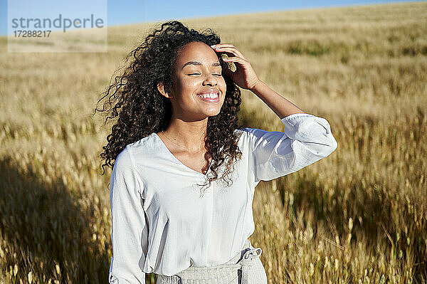 Lächelnde  lockig behaarte Frau mit geschlossenen Augen in einem Weizenfeld an einem sonnigen Tag