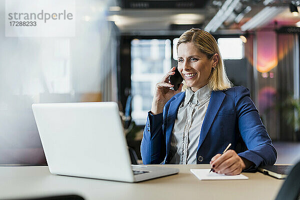 Lächelnde Unternehmerin  die mit einem Smartphone spricht  während sie mit einem Laptop am Schreibtisch sitzt