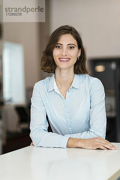 Schöne junge Geschäftsfrau mit braunen Haaren lehnt sich an den Schreibtisch und steht im Büro