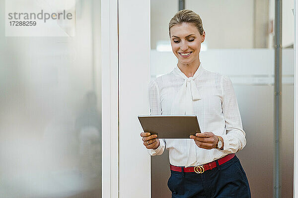 Lächelnde Unternehmerin  die ein digitales Tablet benutzt  während sie an der Tür im Büro lehnt