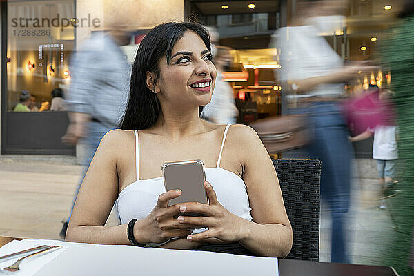 Junge Frau mit Mobiltelefon träumt in einem Straßencafé