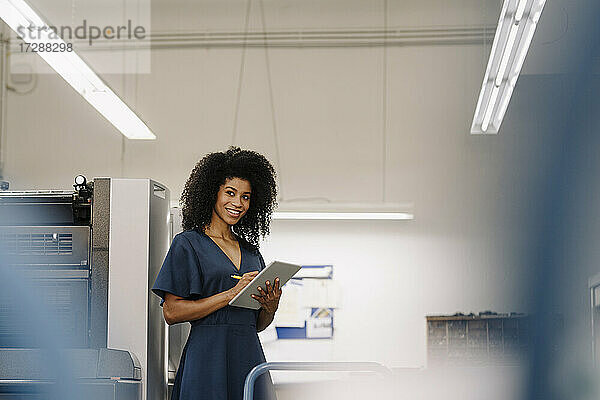 Lächelnde Afro-Unternehmerin  die bei der Arbeit an einem digitalen Tablet in der Industrie wegschaut