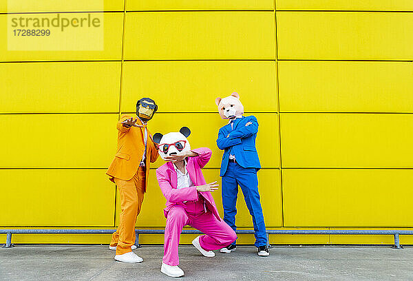 Drei Erwachsene in farbenfrohen Anzügen und Tiermasken posieren gemeinsam vor einer gelben Wand
