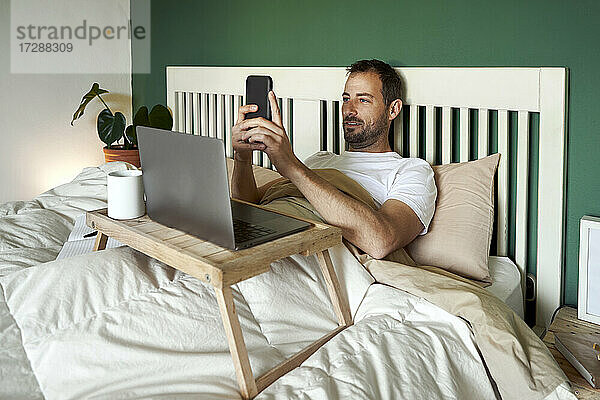 Mann  der ein Mobiltelefon benutzt  während er zu Hause mit seinem Laptop auf dem Tisch liegt