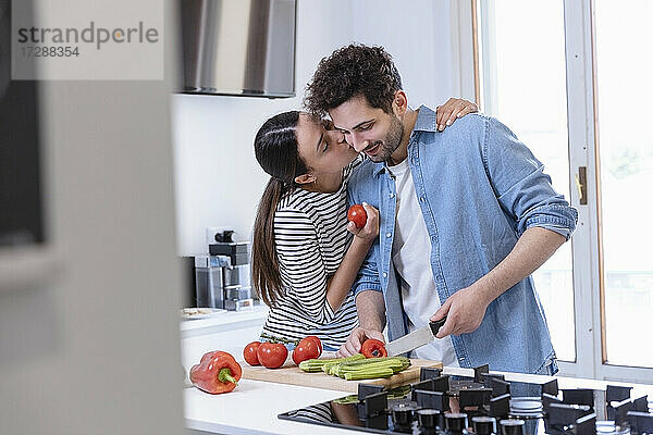 Junge Frau küsst ihren Freund auf die Wange  während sie in der heimischen Küche Paprika schneidet
